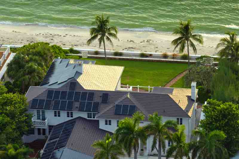 Modern house with rooftop solar panels, surrounded by palm trees along a beachside road.